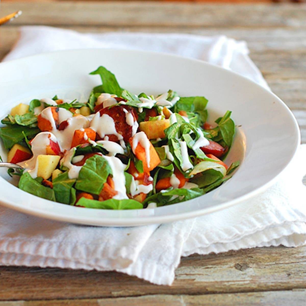 Harvest salad with sweet potato, spinach, turkey kielbasa, bacon, apples, and topped with a creamy homemade maple dressing in a white dish.